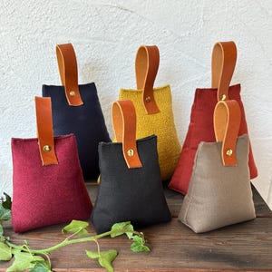 Könnte beinhalten: Sechs pyramidenförmige Türstopper aus Stoff in verschiedenen Farben wie Bordeaux, Marineblau, Gelb, Schwarz, Rot und Beige. Jeder Türstopper hat einen strukturierten Stoffkörper, einen beigen Ledergriff und einen goldenen Metallniet.