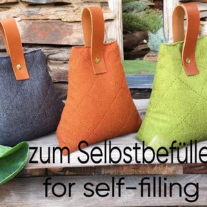Può includere: Tre sacchetti di stoffa grigi, arancioni e verdi, ciascuno con un motivo a rombi e un manico in pelle marrone. Il testo "zum Selbstbefüllen for self-filling" è visibile. Questi sacchetti sono da riempire.