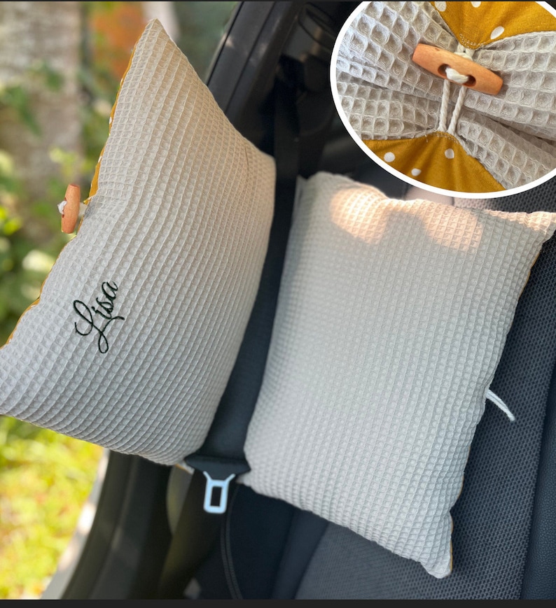 Puede incluir: Dos almohadas para reposacabezas de asiento de coche de tejido de gofre gris claro. Una almohada tiene el nombre "Lisa" bordado en escritura negra. Las almohadas tienen un forro amarillo con lunares y cierre de bot&oacute;n de madera.