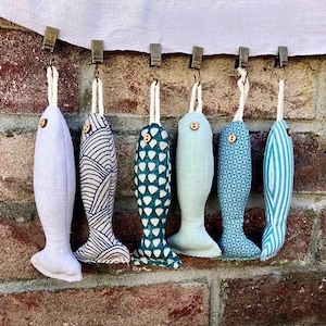Peut inclure: Sept poissons en tissu suspendus à des pinces sur un mur en briques. Les poissons sont de différentes nuances de bleu et de vert avec des motifs différents. Un poisson est blanc.