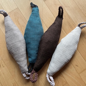 Op de afbeelding: Vier langwerpige, ovaalvormige stoffen zakjes met een textuurweefsel, elk voorzien van een gevlochten lus aan de boven- en onderkant. De zakjes zijn lichtgrijs, talingblauw, donkerbruin en gebroken wit van kleur en staan naast elkaar opgesteld.