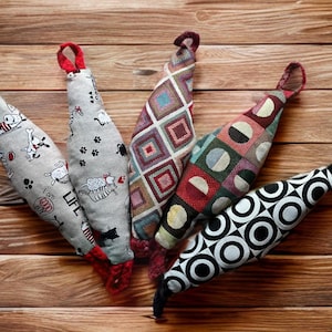 May include: Five dog toys with loop handles in various patterns are displayed on a wooden surface. The toys feature cartoon dogs, paw prints, and geometric designs. Each toy has a red or black loop for easy handling.