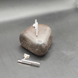 Op de afbeelding: Een paar zilveren oorbelletjes met een geometrisch ontwerp. Elke oorbel heeft drie dunne, rechte staafjes die verticaal zijn gerangschikt.