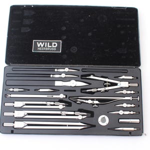 May include: A black hinged case containing a set of silver metal drafting instruments, including various compasses, dividers, and attachments. The interior lid features a black label with the white text WILD HEERBRUGG.