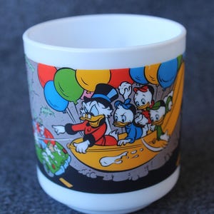 Könnte beinhalten: Ein weißer Becher mit einer bunten Grafik von Dagobert Duck und seinen drei Neffen in einem gelben Boot. Die Szene zeigt leuchtend rote, blaue, grüne und gelbe Luftballons, die über den Figuren vor einem grauen Hintergrund schweben.