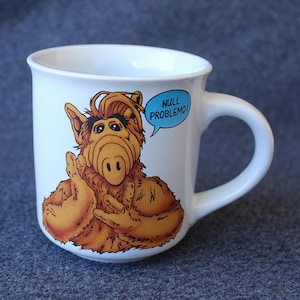 May include: A white ceramic mug featuring a graphic illustration of the character ALF with brown fur, a large nose, and dark eyebrows. A light blue speech bubble next to the character contains the text NULL PROBLEMO in bold black capital letters.