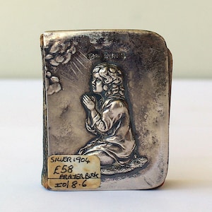May include: Antique silver prayer book, featuring a detailed relief of a person kneeling in prayer, with clouds and light rays. The book is marked "Silver 1904".