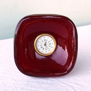 May include: A burgundy-colored, square-shaped clock with rounded corners. The clock face is round with a gold-colored bezel, featuring Roman numerals and minute markers. The clock has a vintage aesthetic.
