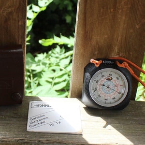 May include: A black altimeter with an orange strap and a white face with black markings. The altimeter reads up to 6000 meters. The altimeter is sitting on a wooden surface with a brown leather case and a small white pamphlet with the word "Garantie" printed on it.