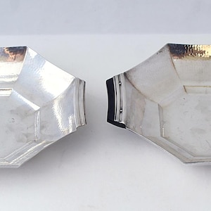 May include: Two octagonal silver-toned bowls with a polished finish. Each bowl features a wide, shallow basin with angled sides and a flat base. The bowl on the right includes two black, curved handles attached to the sides with small metal rivets.