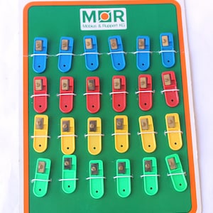 May include: A retail display card featuring twenty-four small pencil sharpeners in blue, red, yellow, and green. Each sharpener has a metal blade and is secured to the green cardboard backing with white thread. The logo M+R Mobius & Ruppert KG is printed at the top.