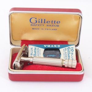 Könnte beinhalten: Ein silberfarbener Sicherheitsrasierer aus Metall mit strukturiertem Griff in einem roten rechteckigen Etui mit goldfarbenem Innenraum. Der Deckel trägt den Schriftzug Gillette Safety Razor Made in England sowie eine blaue Schachtel mit 10 Klingen.