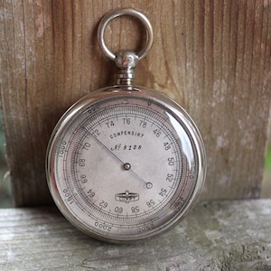 May include: A vintage silver-toned barometer with a clear glass face. The face displays numbers and markings, including the text "COMPENSIRT" and "No 9138." A needle points to a specific measurement. The barometer has a ring at the top for hanging.