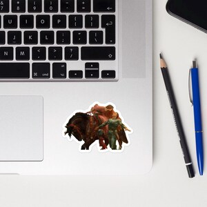 May include: A sticker featuring a fantasy scene of a dark brown horse with riders. The riders are depicted in shades of red and green. The sticker is placed on a laptop.