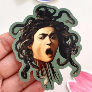 Peut inclure: Un autocollant représentant la tête coupée de Méduse, tirée d'une peinture classique. L'image montre une femme avec des serpents en guise de cheveux, une expression choquée et du sang qui coule de son cou. L'autocollant a une bordure verte.