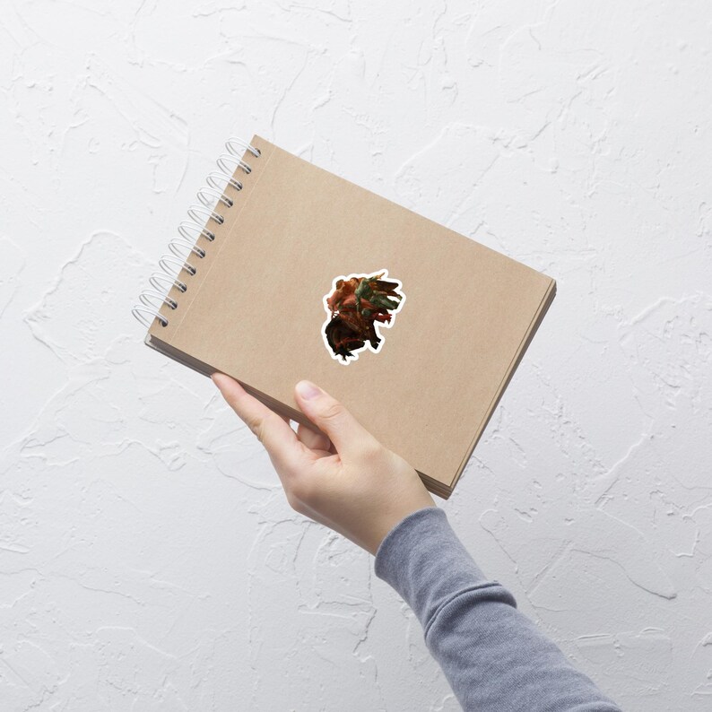 May include: A sticker featuring a detailed illustration of a creature with a mix of red, brown, and green tones. The sticker is affixed to a brown spiral-bound notebook, held by a person.