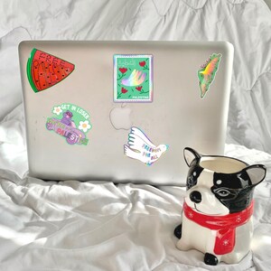 May include: A silver laptop covered in stickers, including a watermelon slice with the word "FREE", a stamp with a hand and the word "PALESTINE", and a dove with the words "FREEDOM FOR ALL". A ceramic dog-shaped planter with a red scarf is in the foreground.