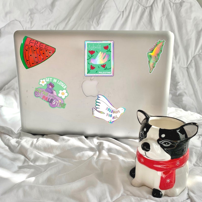 May include: A silver laptop covered in stickers, including a watermelon slice with the word "FREE", a stamp with a hand and the word "PALESTINE", and a dove with the words "FREEDOM FOR ALL". A ceramic dog-shaped planter with a red scarf is in the foreground.