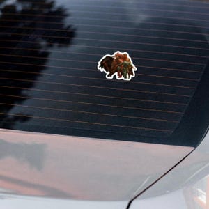 May include: A sticker featuring a detailed illustration of a bison with a person riding on its back. The sticker has a white border and is affixed to a car window. The bison is brown and the rider is wearing green and brown clothing.
