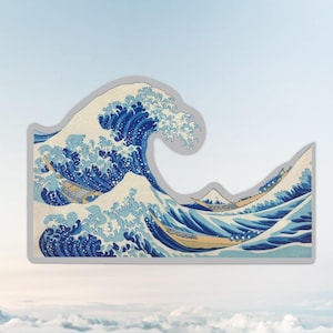 May include: A sticker depicting a large ocean wave with boats, in shades of blue and white. The design is reminiscent of Japanese woodblock prints, with a grey border.