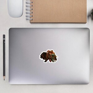 May include: A sticker featuring a fantasy scene with a large, dark brown creature, possibly a bull or bison, ridden by figures in red and green attire. The sticker is on a gray surface.
