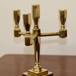 Könnte beinhalten: Ein goldfarbener Metall-Kandelaber mit fünf Kerzenhaltern. Der Kandelaber hat einen quadratischen Sockel und einen zentralen Schaft mit vier Armen, die sich nach außen erstrecken. Jeder Arm hat einen dekorativen, achteckigen Kerzenhalter.