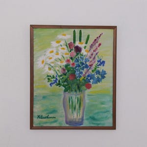 Pode incluir: Pintura emoldurada de um arranjo floral num vaso de vidro. O buquê apresenta margaridas brancas, flores azuis, botões vermelhos e detalhes roxos. O fundo é uma mistura de tons verdes e amarelos, com uma moldura marrom.