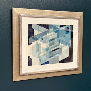 Könnte beinhalten: Abstrakter geometrischer Kunstdruck in einem silbernen Rahmen. Das Kunstwerk zeigt sich überlappende blaue und weiße Formen auf dunklem Hintergrund. Der Rahmen hat eine silberne Oberfläche und ist an einer dunkelblauen Wand befestigt.