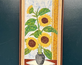 May include: A framed painting of sunflowers in a vase. The sunflowers have yellow petals and brown centers, with green leaves. The vase is black and sits on a white tablecloth. The frame is brown with a woven texture.