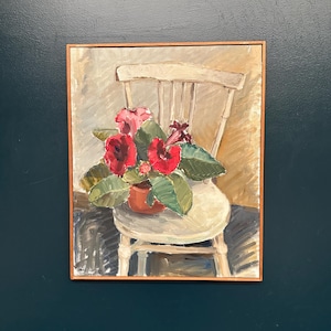 Puede incluir: Una pintura al óleo en un marco de madera que representa una planta en maceta con flores rojas vibrantes y grandes hojas verdes sobre una silla de madera clara. El fondo presenta tonos beige neutros y la firma del artista es visible en la esquina.