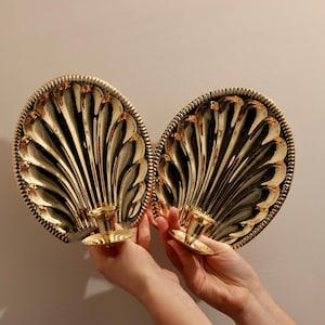 Könnte beinhalten: Zwei goldfarbene, muschelförmige Wandleuchter. Jeder Leuchter hat ein detailliertes, fächerartiges Design mit einem gewellten Rand und einem Kerzenhalter. Die Leuchter werden von Händen vor einem neutralen Hintergrund gehalten.