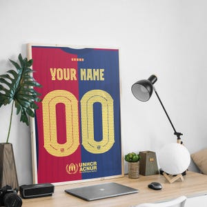 May include: A framed poster of a soccer jersey with a blue and red design. The jersey has a yellow number "00" and the text "YOUR NAME" on the front. The poster also includes the UNHCR logo and the text "UNHCR ACNUR The UN Refugee Agency".