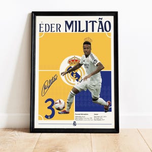 May include: A yellow and blue poster with a photo of a soccer player wearing a white jersey with the number 3 on it. The player is wearing black shorts and white socks. The poster has the text "ÉDER MILITÃO" at the top and the player's signature at the bottom. The poster also includes the player's personal information and career information.