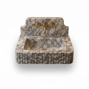 May include: A wall-mounted sink crafted from white marble with prominent grey veining. The basin features a rectangular shape with a fluted front edge and an integrated backsplash. It is equipped with a central brass faucet and two brass handles.