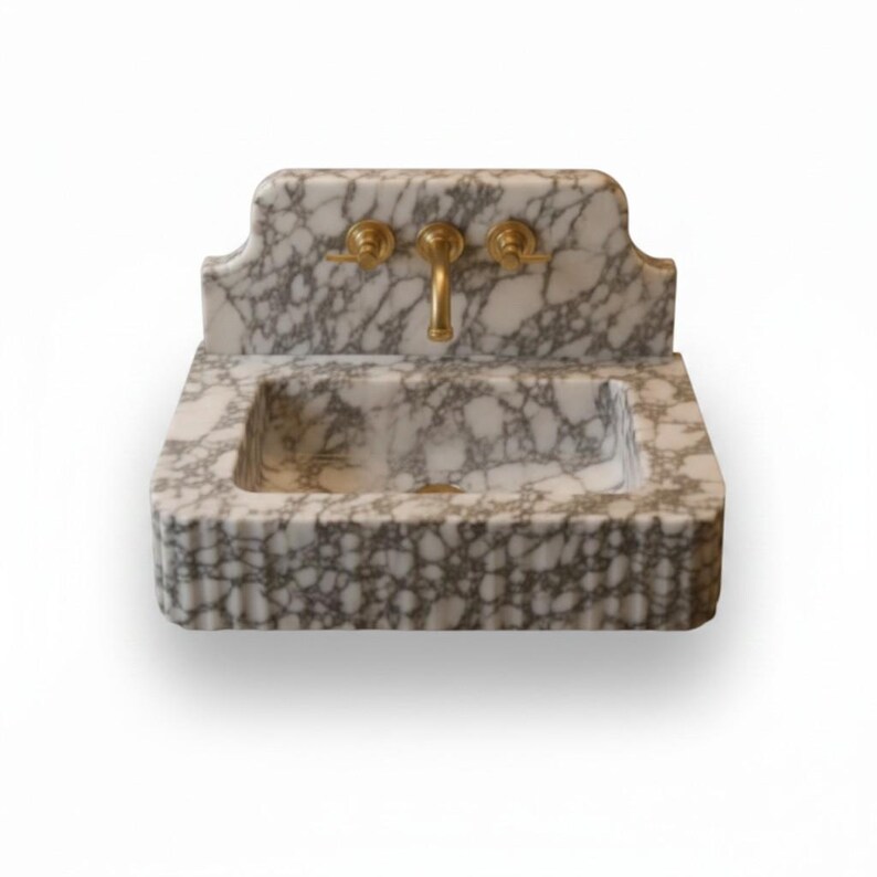 May include: A wall-mounted sink crafted from white marble with prominent grey veining. The basin features a rectangular shape with a fluted front edge and an integrated backsplash. It is equipped with a central brass faucet and two brass handles.