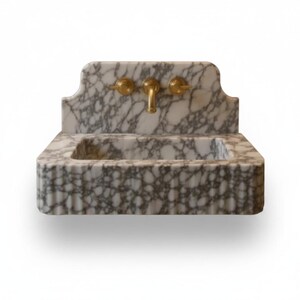 May include: A wall-mounted bathroom sink crafted from white marble with prominent grey veining. The rectangular basin features a fluted front edge and an integrated backsplash. It is equipped with a central brass faucet and two brass handles.