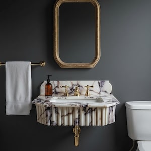 May include: A wall-mounted white marble sink with purple veining and a fluted front, featuring gold-toned faucets and a matching gold-toned drain pipe. Above the sink hangs a rectangular mirror with a decorative gold-toned frame.