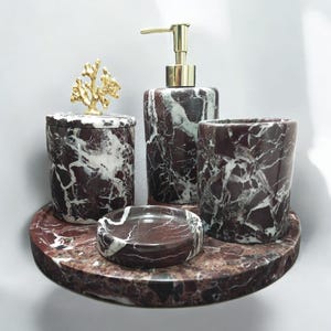 May include: A set of four bathroom accessories made of brown and white marble. The set includes a soap dispenser, a toothbrush holder, a jar with a gold coral lid, and a soap dish. The accessories are arranged on a round marble tray.