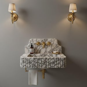 May include: A wall-mounted white and grey marble sink featuring a fluted front, brass faucet, and two matching brass handles. A soap dispenser and a small dish with soap sit on the basin. A white towel hangs from a brass bar below the sink.