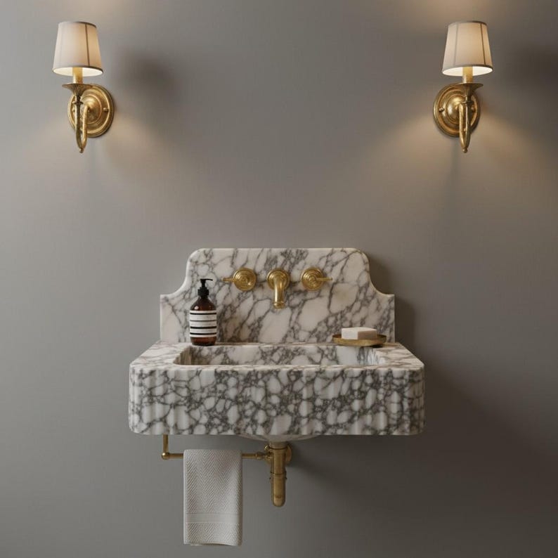 May include: A wall-mounted white and grey marble sink featuring a fluted front, brass faucet, and two matching brass handles. A soap dispenser and a small dish with soap sit on the basin. A white towel hangs from a brass bar below the sink.