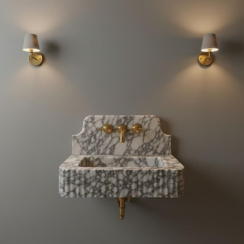 May include: A wall-mounted white and grey marble sink featuring a rectangular basin with a fluted front edge. The sink includes a matching marble backsplash and a brass faucet with two matching brass handles. Two wall sconces flank the sink.