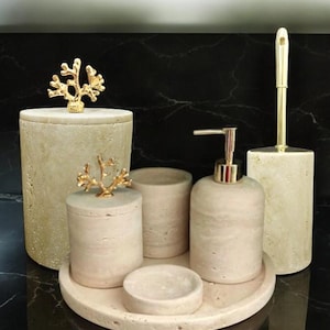 Könnte beinhalten: Ein Set Badezimmerzubehör aus beigefarbenem Beton mit goldenen Akzenten. Das Set beinhaltet einen Seifenspender, einen Zahnbürstenhalter, ein Tablett und ein kleines dekoratives Glas. Das Zubehör ist auf einer schwarzen Marmorfläche angeordnet.