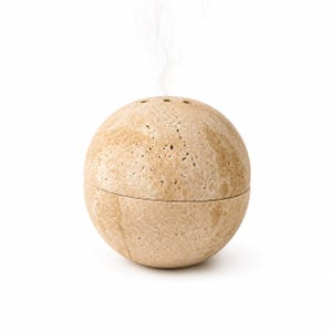 May include: A spherical stone diffuser with a porous, beige, and tan textured surface. The object is divided into two horizontal sections and features three small circular openings at the top, from which a thin stream of white vapor is rising.