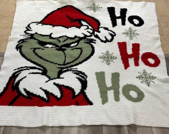 May include: A white crocheted blanket with a green Grinch wearing a red Santa hat and the words "Ho Ho Ho" in black and red.
