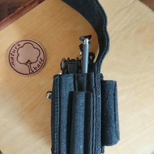 Op de afbeelding: Zwarte nylon tas met een ritssluiting en meerdere vakken. De tas is ontworpen om een multitool en andere kleine items te bevatten. De tas heeft een drukknopsluiting aan de bovenkant en een lederen patch met de tekst "nature base" aan de voorkant.