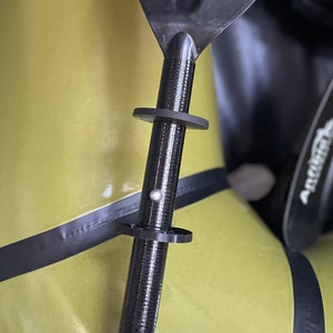 Könnte beinhalten: Nahaufnahme eines schwarzen Paddels mit einem silbernen Knopf und zwei schwarzen Ringen. Das Paddel ist an einem gelben Schlauchboot befestigt.
