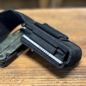 Könnte beinhalten: Eine schwarze Nylon-Multitool-Scheide mit einer silbernen Metallfeile. Die Scheide ist für ein Multitool konzipiert, wobei ein Multitool mit Tarnmuster teilweise sichtbar ist. Die Scheide hat eine Gürtelschlaufe.