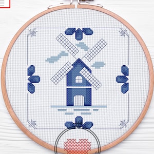 Può includere: Un motivo a punto croce blu e bianco di un mulino a vento con nuvole e acqua. Il motivo è incorniciato in un telaio da ricamo in legno.