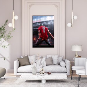 Op de afbeelding: Een witte bank met bruine kussens staat in een woonkamer met een witte lamp, een kleine tafel en twee witte stoelen. Een ingelijst poster van een voetballer in een rood shirt met nummer 7 op de rug hangt aan de muur.