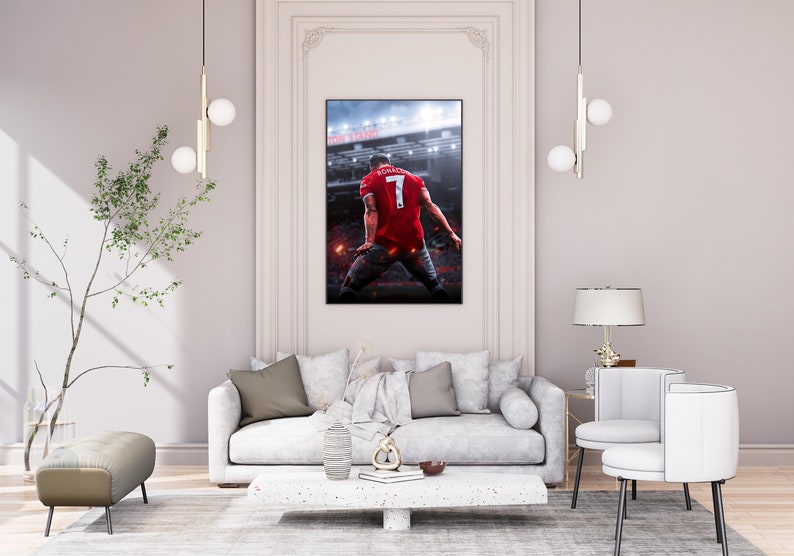 Op de afbeelding: Een witte bank met bruine kussens staat in een woonkamer met een witte lamp, een kleine tafel en twee witte stoelen. Een ingelijst poster van een voetballer in een rood shirt met nummer 7 op de rug hangt aan de muur.